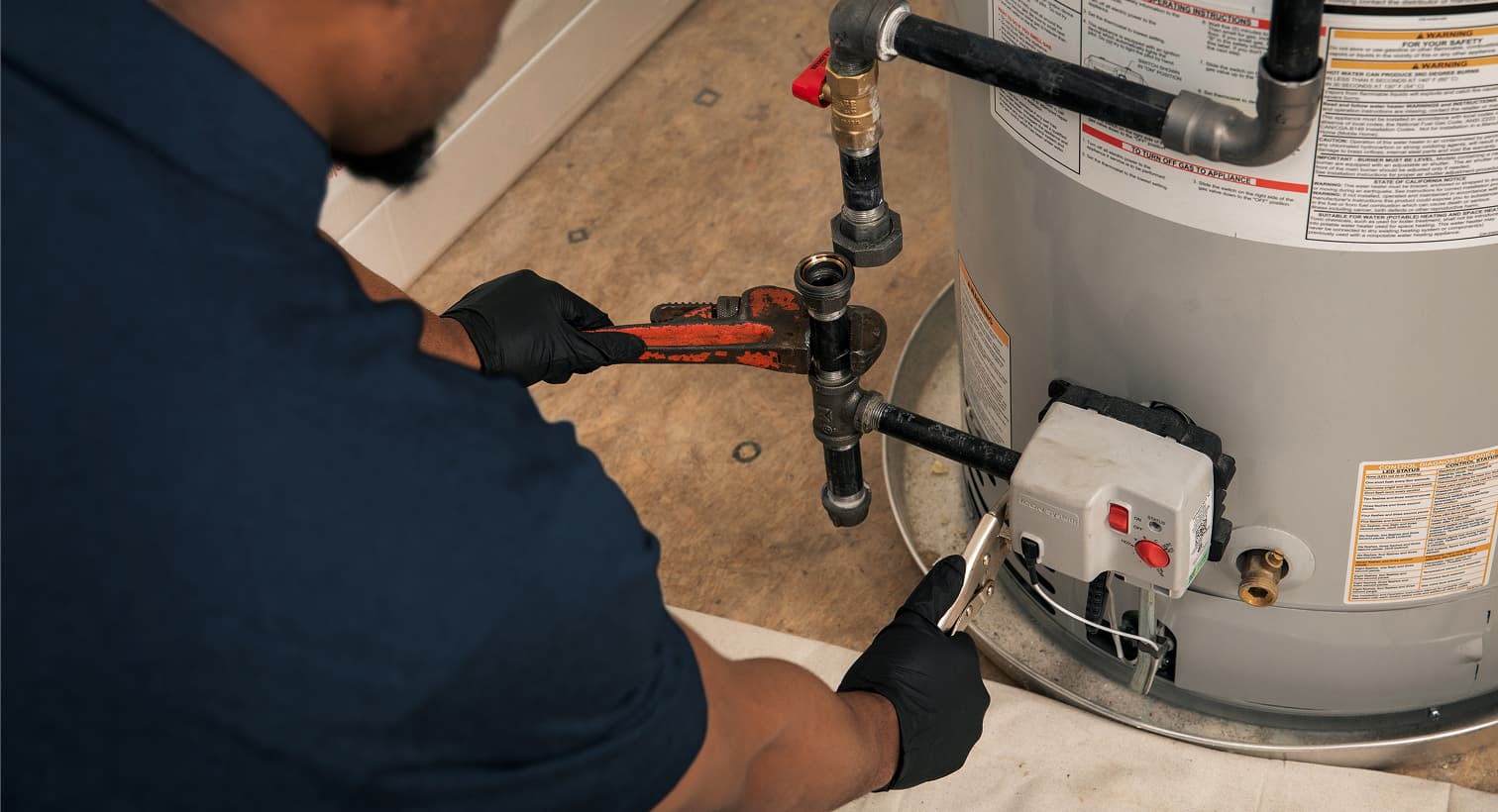 A Fenwick Waychoff's plumber wearing black gloves uses a pipe wrench to repair the gas line and control valve assembly on a residential water heater tank.
