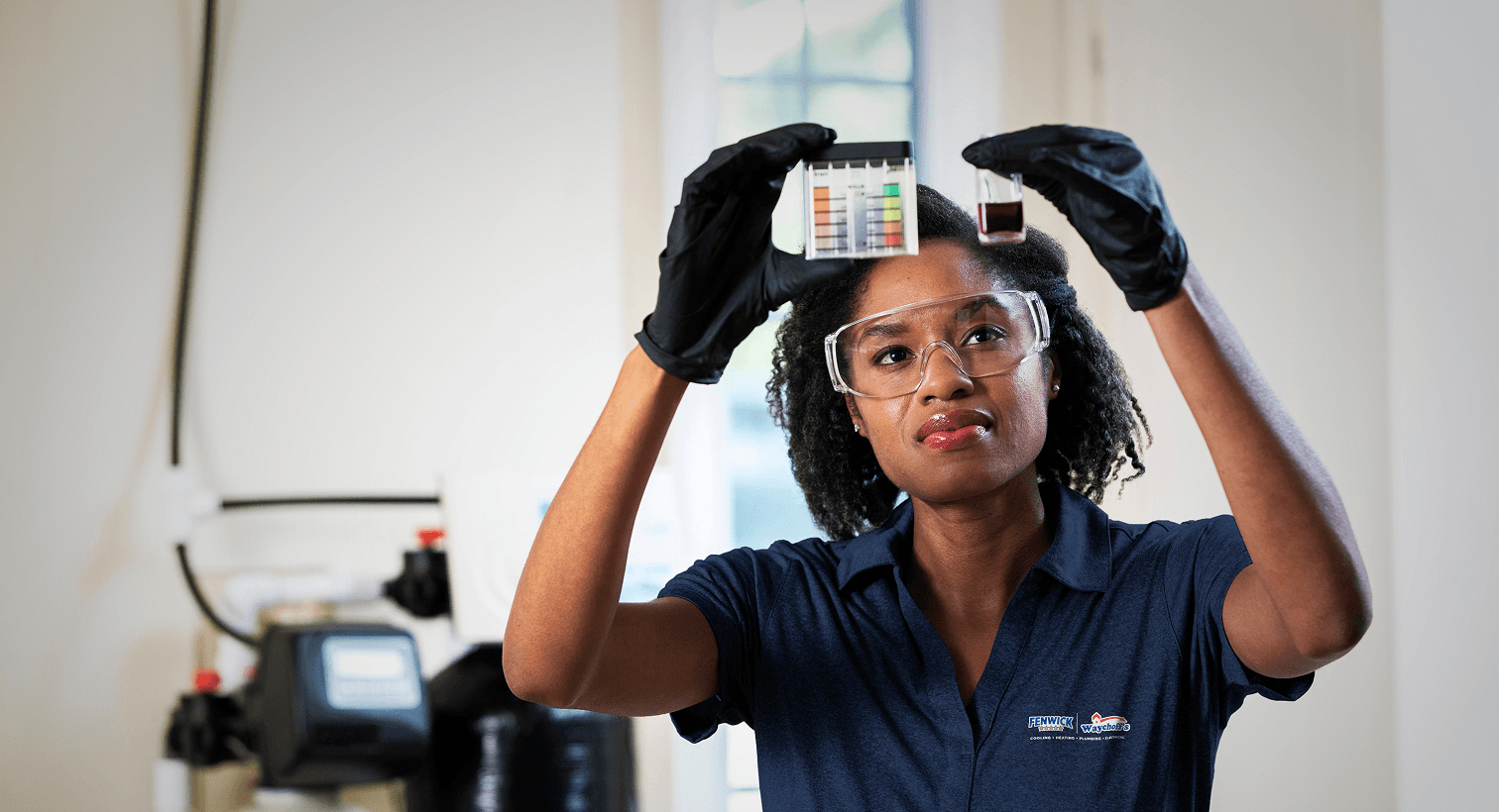 A Fenwick technician in a blue uniform and safety gear compares a water sample to a testing chart in front of a water softener system.